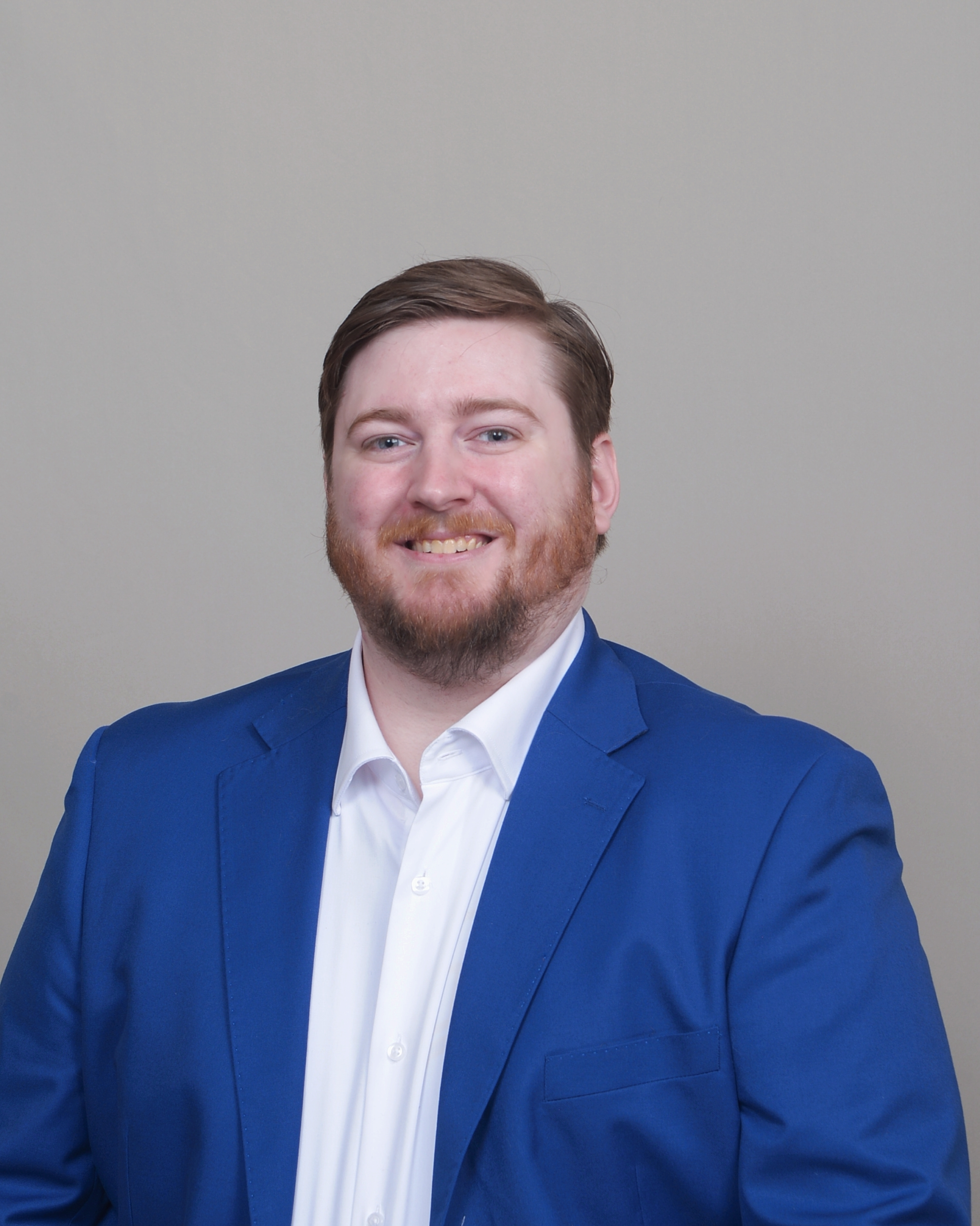 A headshot above the waist, facing slightly left and smiling. With a blue suit jacked and white dress shirt with no tie. With a short beard, and parted brown hair to the side.